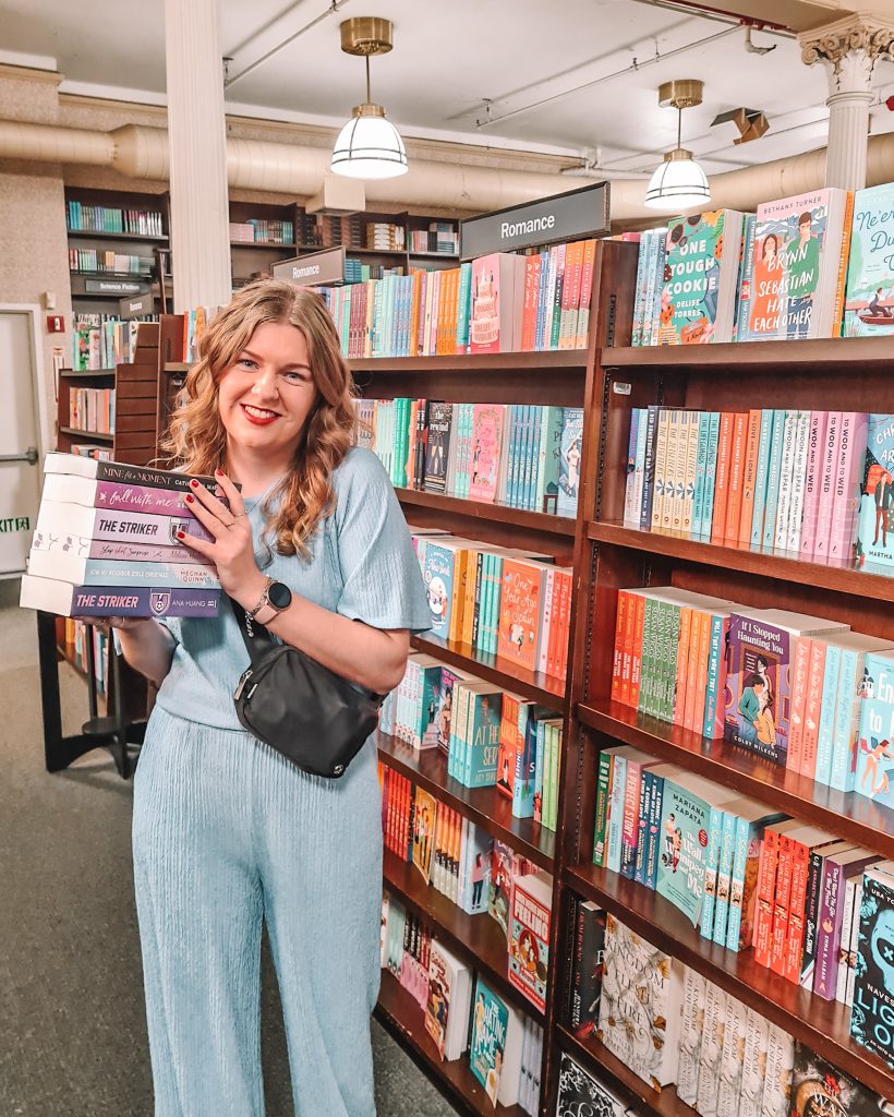 Charlotte stands in front of a romance bookshelf in a bookstore, holding a stack of six books. She's wearing a blue top and pants and has a small black bag slung over her shoulder.
