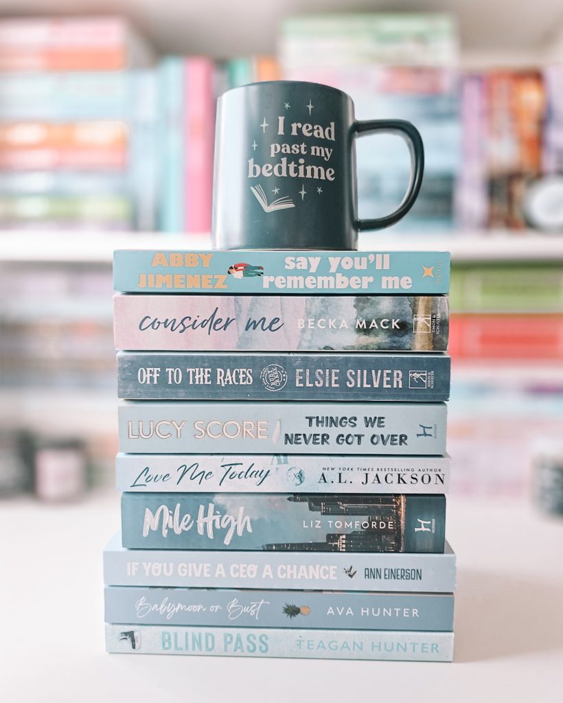 A blue mug reading "I read past my bedtime" sitting atop a stack of books facing spine out, each with a blue color scheme, in front of an out of focus bookshelf.