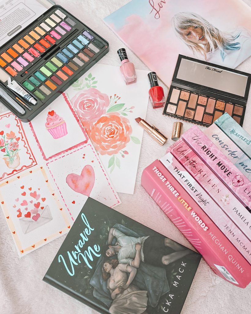 Flatlay featuring Unravel Me by Becka Mack laid cover-up alongside a stack of six other books with the spines facing out. The books are arranged alongside watercolor paintings of roses, a cupcake, an envelope with hearts coming out, and a large heart, as well as an open watercolor set, a neutral-toned makeup palette and tube of brown lipstick, red and pink bottles of nail polish, and a vinyl record of the album Lover by Taylor Swift.