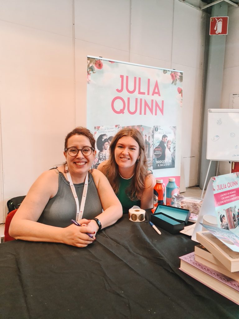 Julia Quinn, a light-skinned woman with brown hair tied back and round glasses, sits at a table holding a pen as she smiles at the camera. Charlotte of @charlottes.loveforbooks crouches down beside her. On the table are copies of Quinn's books, bottles of water, and stickers, and behind her is a large sign with her name on it and photos of her books, along with a whiteboard on an easel.