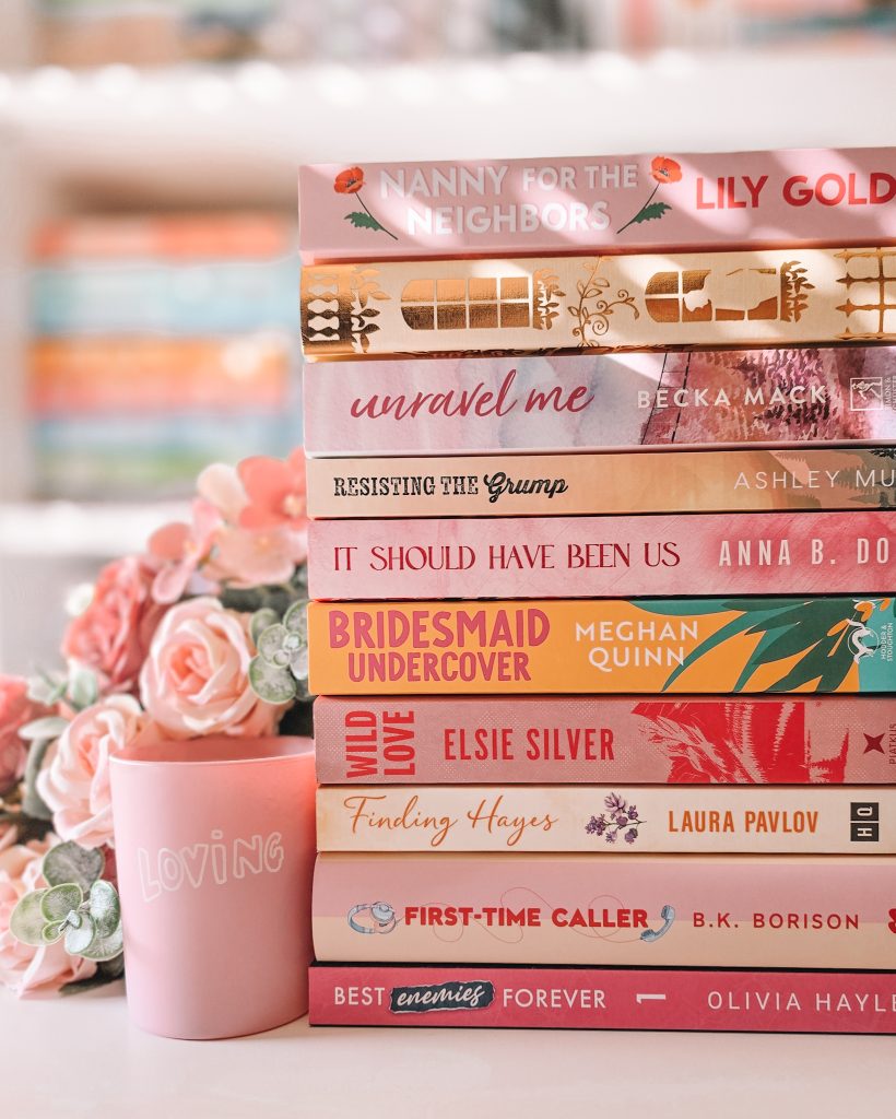 A stack of books, each with a pink color scheme, facing spine out next to a pale pink cup that reads "Loving" and a bunch of pink roses, in front of an out of focus bookshelf.