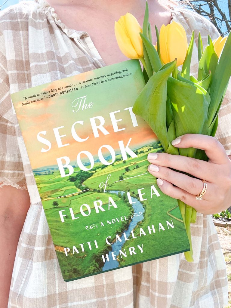 A person in a light plaid dress holds a bouquet of yellow tulips and a copy of The Secret Book of Flora Lea by Patti Callahan Henry. The book’s vibrant green cover features a pastoral landscape with a winding river.