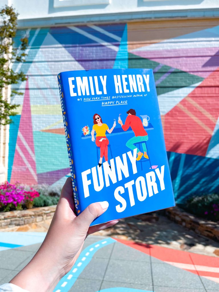 Courtney holds up a copy of Funny Story by Emily Henry against a vibrant, geometric mural painted on a wall. The book’s bright blue cover features illustrated characters clinking drinks at a bar. The background includes colorful triangular shapes in pink, teal, purple, and white.