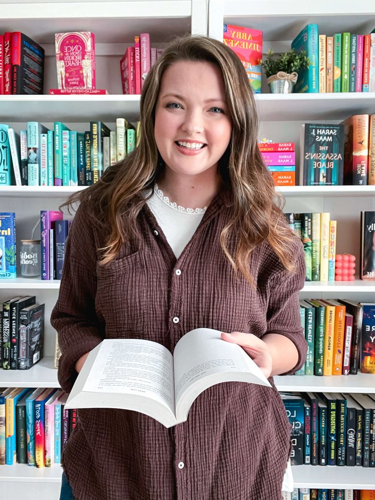 A woman with long brown hair stands smiling in front of a colorful rainbow-organized bookshelf. She wears a brown sweatshirt and holds an open book in her hands, creating a cozy and inviting reading atmosphere.