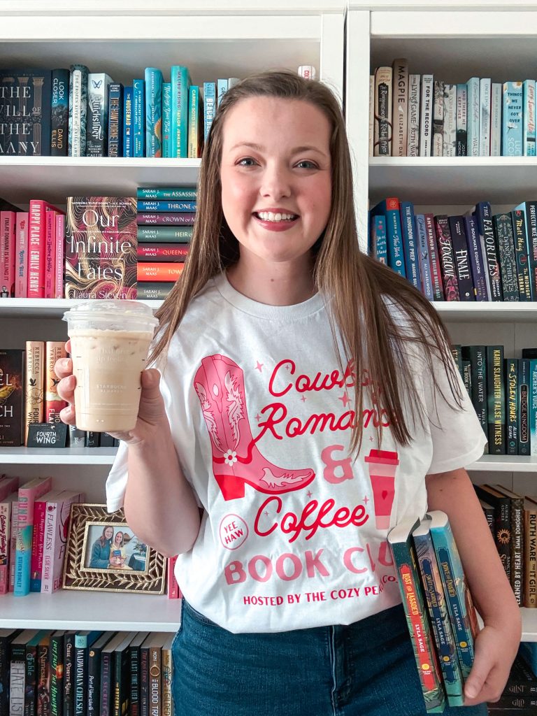 Courtney stands in front of a colorful rainbow-organized bookshelf, holding an iced coffee in one hand and a stack of books in the other. She wears a white graphic t-shirt that reads “Cowboy Romance & Coffee Book Club,” with pink illustrations of a cowboy boot and coffee cup.