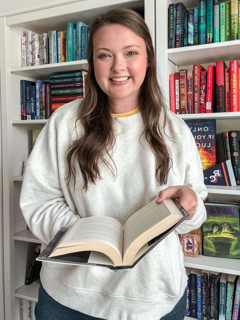 A woman with long brown hair stands smiling in front of a colorful rainbow-organized bookshelf. She wears a light gray sweatshirt and holds an open book in her hands.