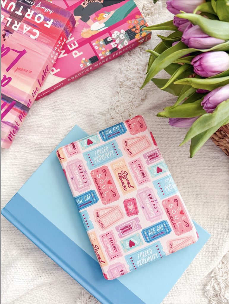 A flat lay of colorful romance-themed items on a soft white blanket. A pastel pink book sleeve with phrases like “Soulmates,” “Second Chance,” and “Forced Proximity” sits on top of a light blue hardcover book. To the left are two vibrant romance novels, including Carly Fortune’s "Meet Me at the Lake" and "Practice Makes Perfect." A bouquet of fresh purple tulips peeks in from the top right.
