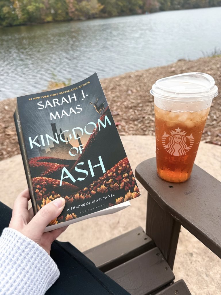 Courtney holds Kingdom of Ash by Sarah J. Maas while sitting by a calm lakeside. The book's dark, autumn-themed cover features a landscape of red and orange leaves. A Starbucks iced drink rests on a wooden armrest next to the book. The background includes peaceful water and trees just beginning to turn for fall.