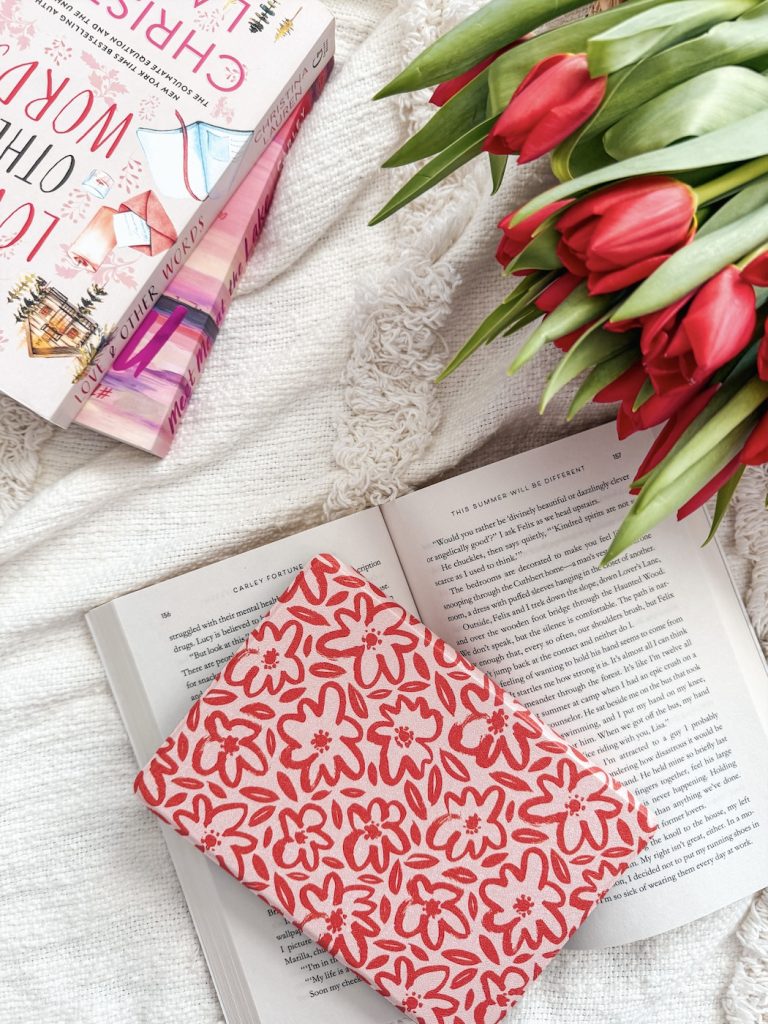 A cozy bookish flat lay featuring a red floral-patterned Kindle sleeve resting on an open book. Nearby are two pastel romance novels by Carley Fortune and Christina Lauren stacked on a cream knit blanket. A vibrant bouquet of red tulips frames the scene.