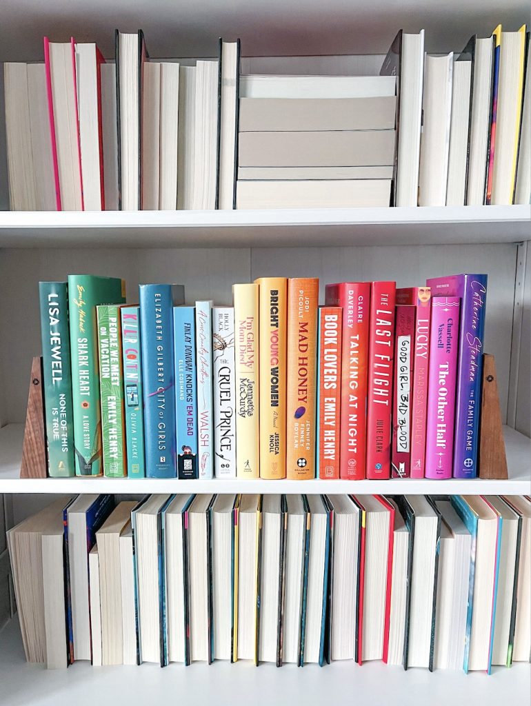 A neatly arranged bookshelf featuring a rainbow gradient of book spines on the middle shelf, ranging from green to pink to purple. The top and bottom shelves show books turned with their pages outward, emphasizing texture and color contrast.