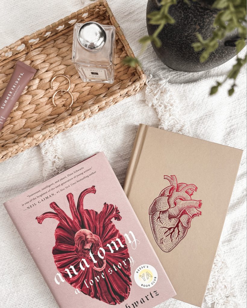 A flat lay featuring the book Anatomy: A Love Story by Dana Schwartz with its illustrated cover beside a matching hardcover edition embossed with an anatomical heart. The setup includes a woven tray holding a bottle of perfume, gold jewelry, and lip balm, placed on a soft cream blanket with a potted plant in view.