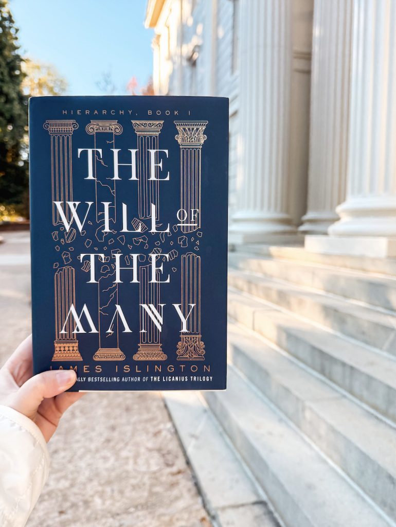 A hand holds the book The Will of the Many by James Islington in front of a grand neoclassical building with tall white columns and stone steps. The book cover features a deep navy background with gold-embossed columns and shattered pieces.