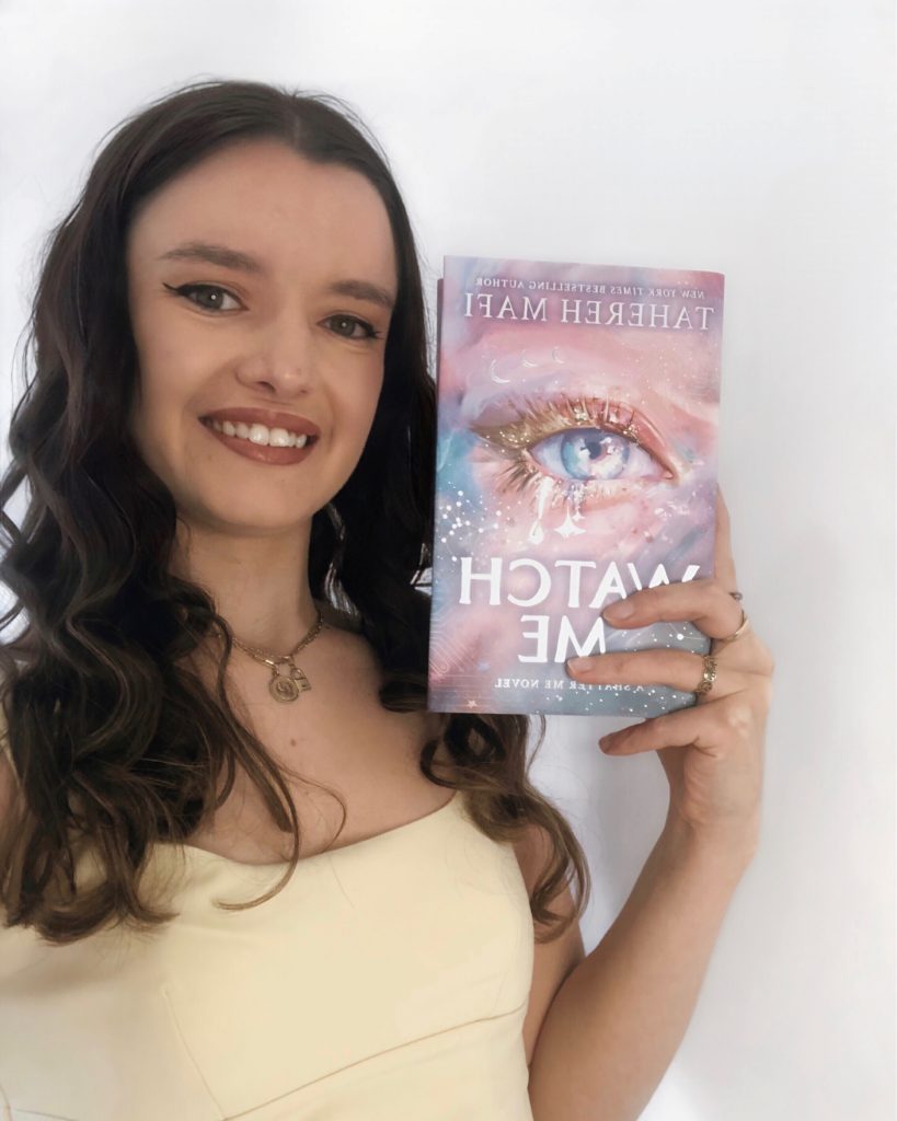 Bookstagrammer Carla in a pale yellow shirt, with her hair perfectly curled, smiling and holding up a hardback cover of Watch Me by Tahereh Mafi. The cover is a sparkling blue eye crying glittery tears.