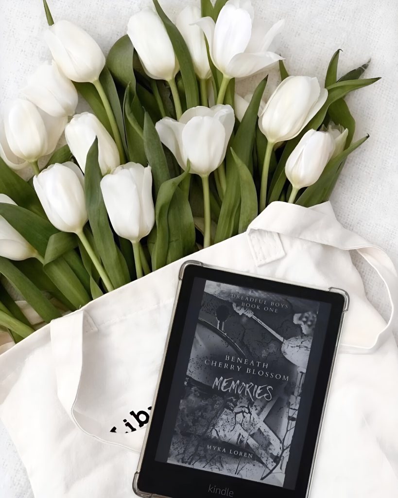 A gorgeous bouquet of white tulips with thick green leaves coming out of a white bag with a kindle resting on top. On the kindle Beneath Cherry Blossom Memories by Myka Loren is displayed.