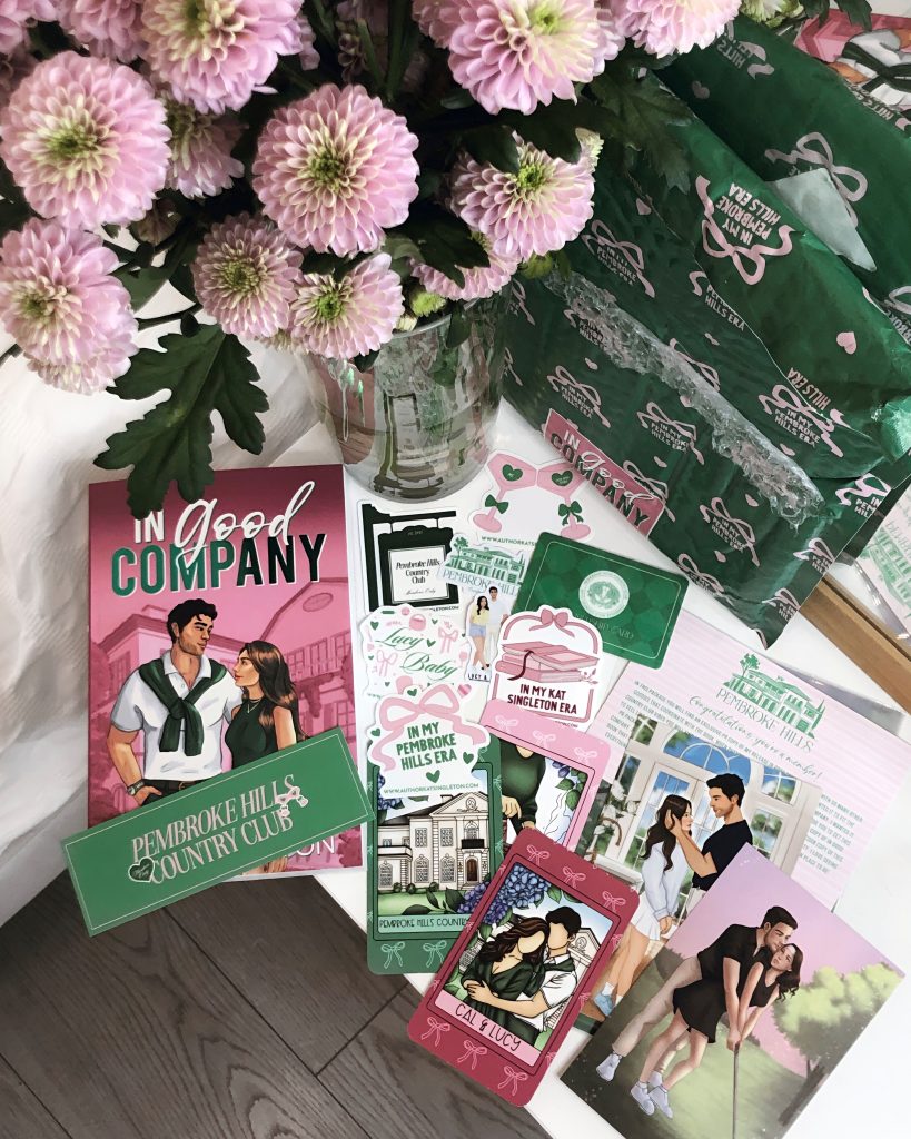 On a white bedside table sits the romance novel In Good Company by Kat Singleton with a pink background surrounding a couple in matching forest green in front of a country club. Behind a vase of purple flowers is the books decorated package that contained the remainder of the contents on the table: multiple stickers, a Pembroke Hills Country Club bookmark, and graphics of the couple from the book.