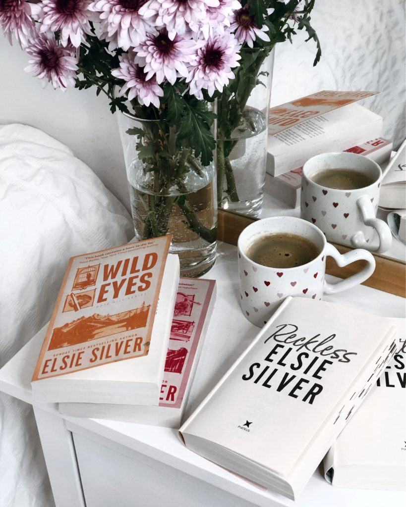 On a white bed side table sits a glass vase with purple flowers, a white mug with red and purple hearts filled with coffee, and multiple Elsie Silver books. The two exposed are Wild Eyes and Reckless.