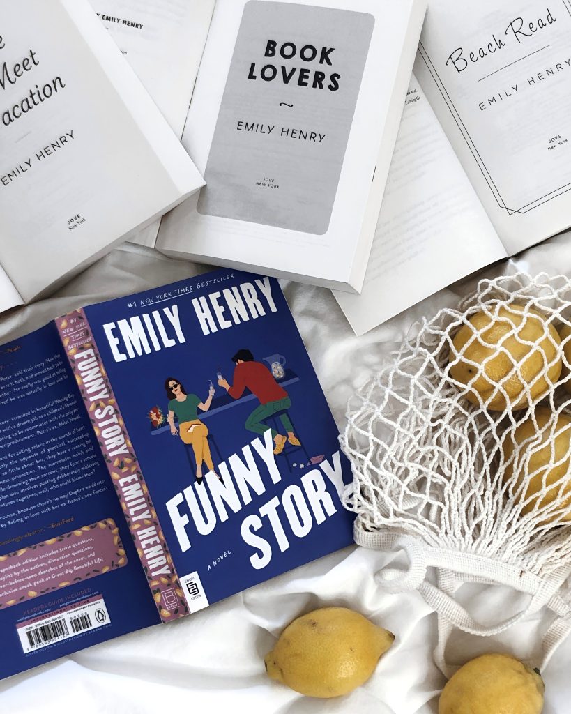 The royal blue romance book cover Funny Story by Emily Henry lays open on a white bed beside multiple other Emily Henry books open to their white title pages. Beside them sits a knit white fruit bag containing lemons.