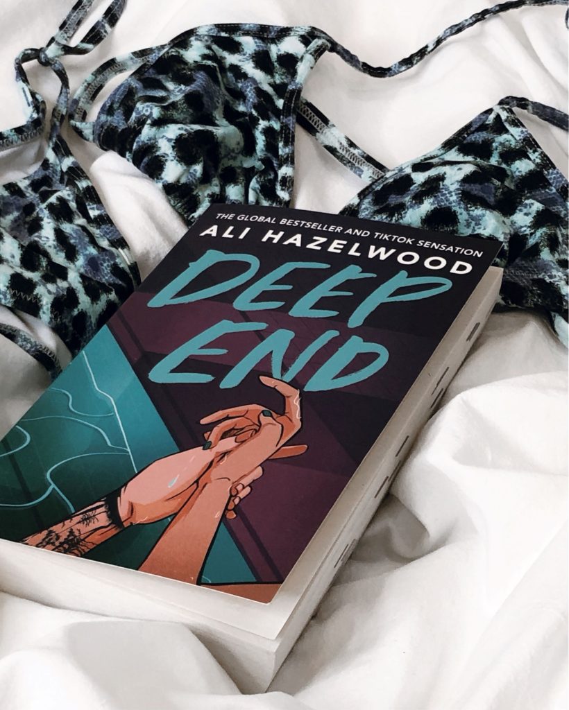 On top of white sheets is a blue cheetah print bikini with the romance book Deep End by Ali Hazelwood on top of it. The cover shows two hands, one with nail polish and the other with tattoos, holding each other.