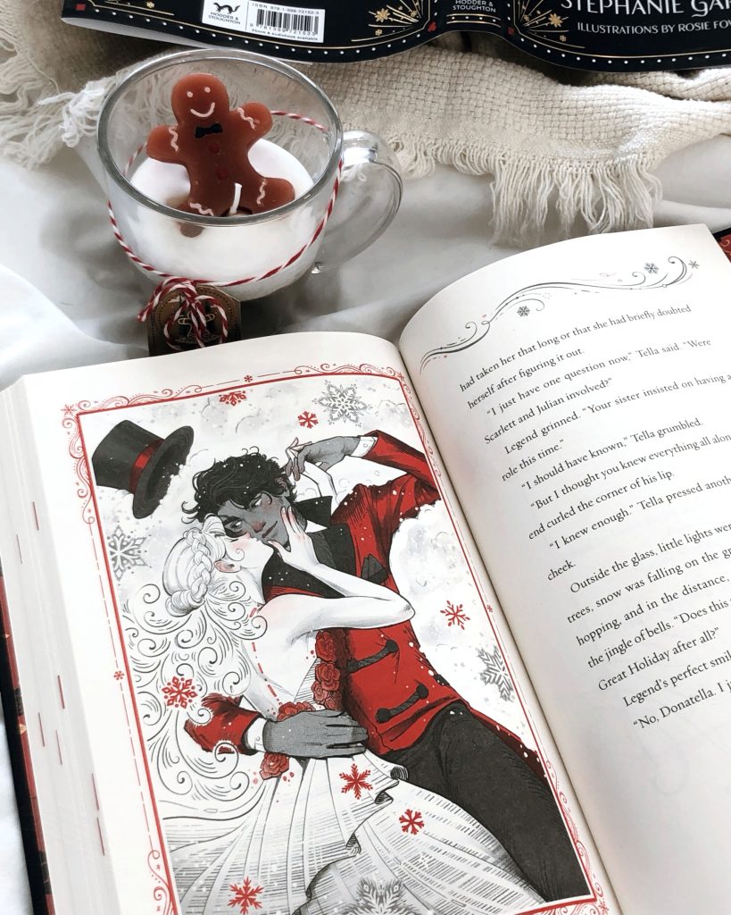 On top of white sheets a fringed white blanket peeks off camera with a gingerbread man candle in a glass jar with red and white twine around it. Below the mug is an open book with an illustrated page of a man in a red formal coat with his black top hat coming off staring at a woman dressed in all white holding his face and kissing his cheek.