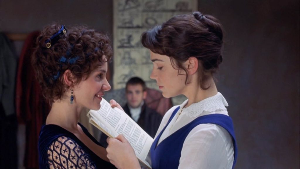 A still from the 1999 movie Mansfield Park. A young woman with blue ribbons woven through her curly bun sneers at another young woman in a blue dress and white blouse who stares down at a book she holds in front of her.