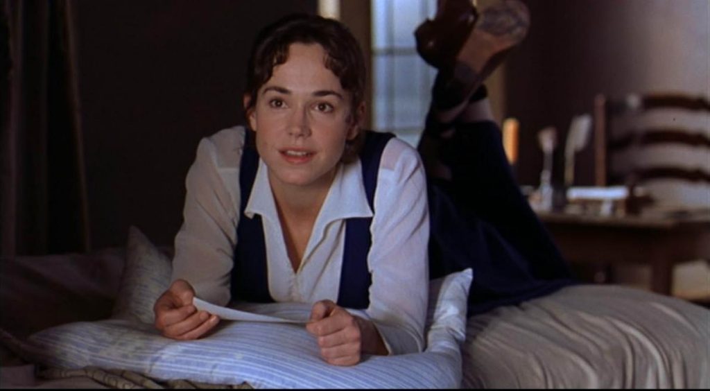 A still from the 1999 movie Mansfield Park. A young woman in a black dress with a white blouse lays casually on a bed with her feet swinging in the air. She holds a letter in her hand and stares wistfully into the distance.