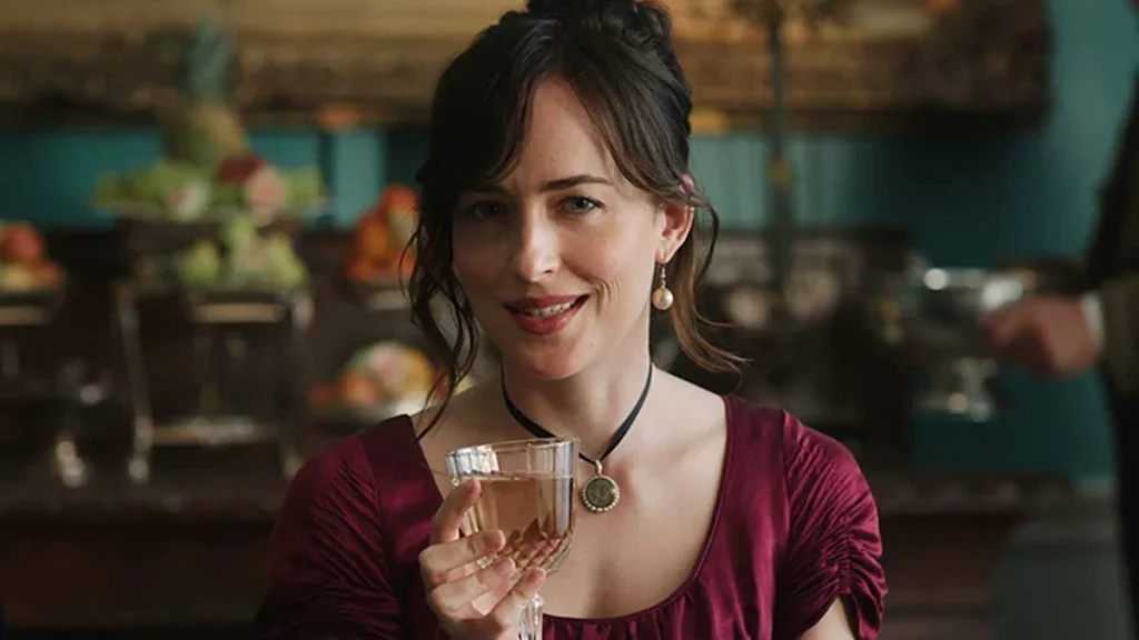 A still from the 2022 movie Persuasion. A young woman in a maroon evening gown holds a glass of white wine and smiles cheekily into the camera. Behind her is a buffet table loaded with food.