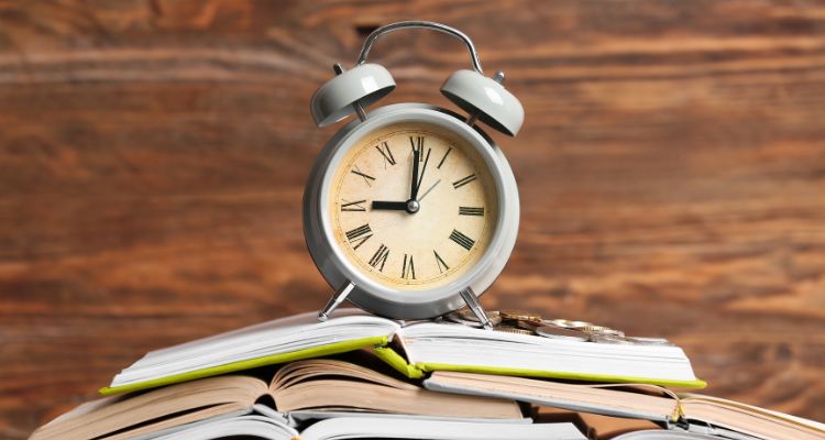 Image depicting an analog alarm clock on top of a stack of open books in front of a wooden background.