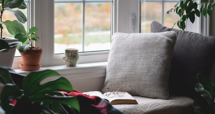 Image depicts a bay window seat with pillows and a book and glasses on the cushion. There is a mug and many plants along the windowsill.