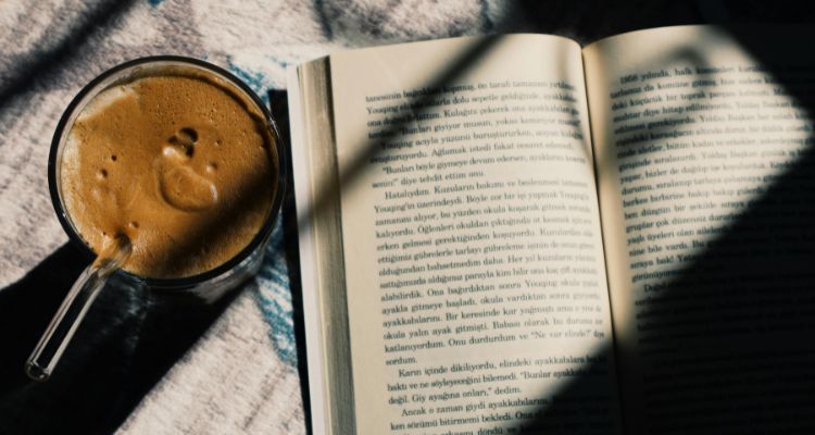 Image depicts an overhead shot of an opened book next to a mug of coffee with sunlight from the window shining on half of the book.