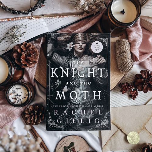 Flatlay featuring a copy of The Knight and the Moth by Rachel Gillig laid atop neutral-colored fabrics, surrounded by journals, candles, berries, pinecones, an envelope, a roll of twine, and open books.
