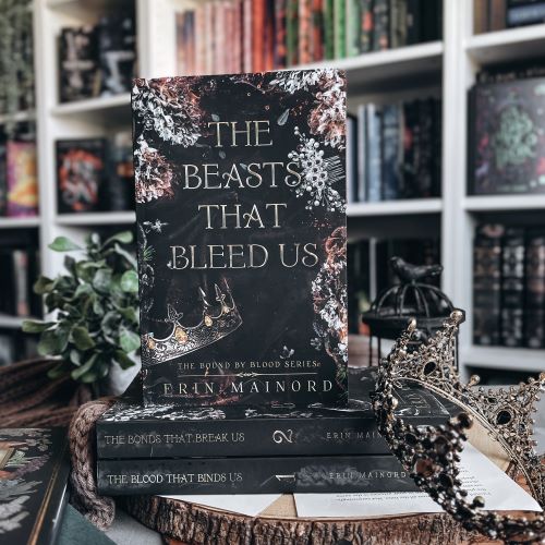 A copy of The Beasts That Bleed Us by Erin Mainord stands atop the first two books in the series, which rest on a tree ring slice in front of a potted plant, black birdcage, and bookcase. A crown sits artfully on the corner of the stack of books.
