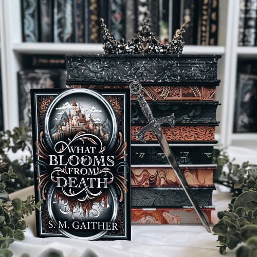 A stack of books with a crown perched on top sits an a rumpled white cloth, surrounded by leaves. In front of the stack of books is a copy of What Blooms from Death by S.M. Gaither, alongside a miniature sword.