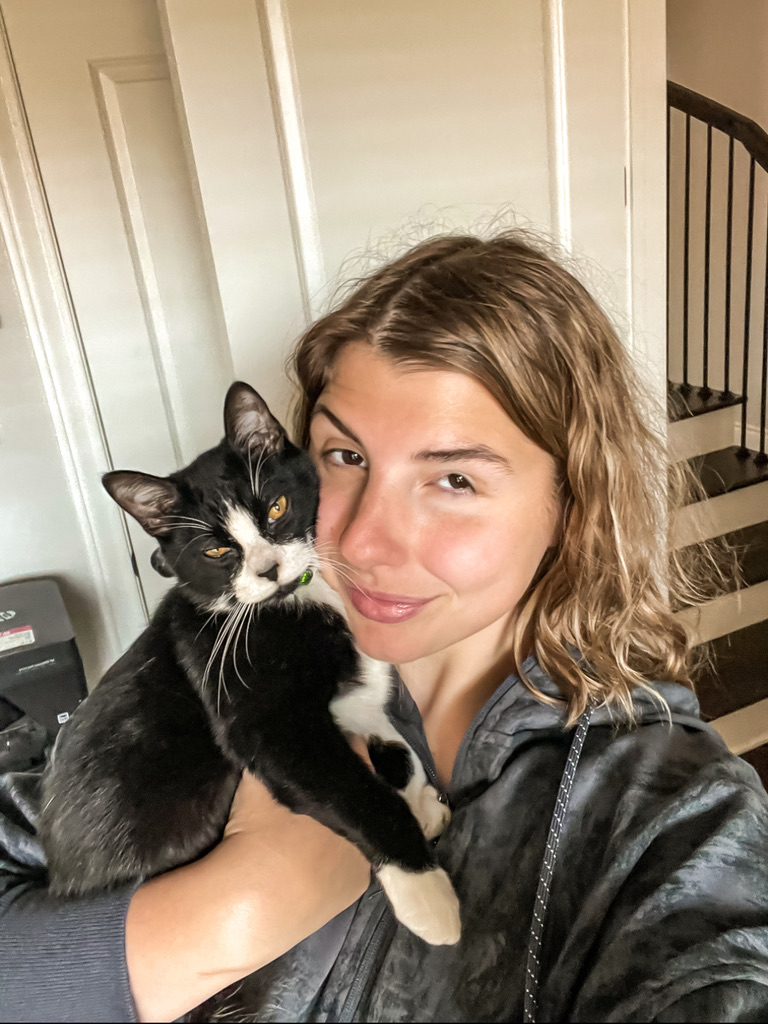 A photo of a woman with shoulder-length dirty-blonde hair holding a tuxedo cat with yellow eyes.