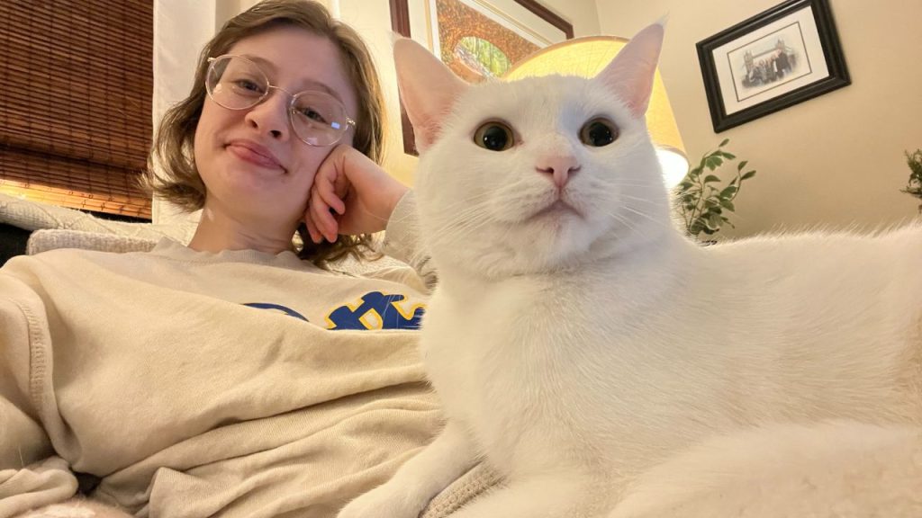 A photo of a woman with short, brown hair and glasses, smiling, and a white cat with big, yellow eyes.