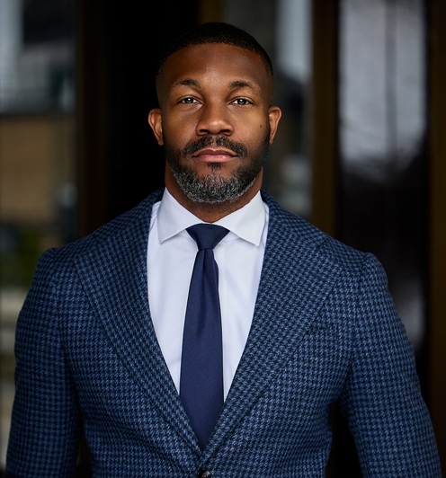 Mayor Randall Woodfin, a black man in a navy blue houndstooth suit and tie with a trim back and grey beard stands powerfully before the camera.