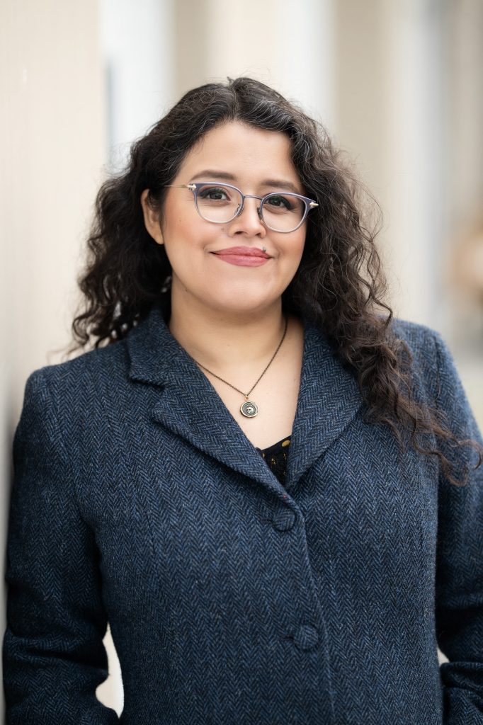 Silvia Moreno-Garcia smiling with with her natural dark waves loose. She wears a pair of gray metallic glasses that complements her blazer.