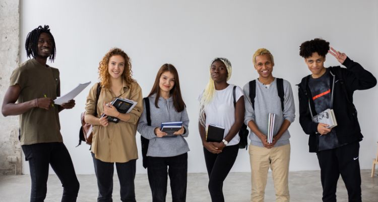 A group of smiling youths diverse in skin tone, hair color/texture, and gender presentation stand in an open room with books in hand and wearing backpacks.