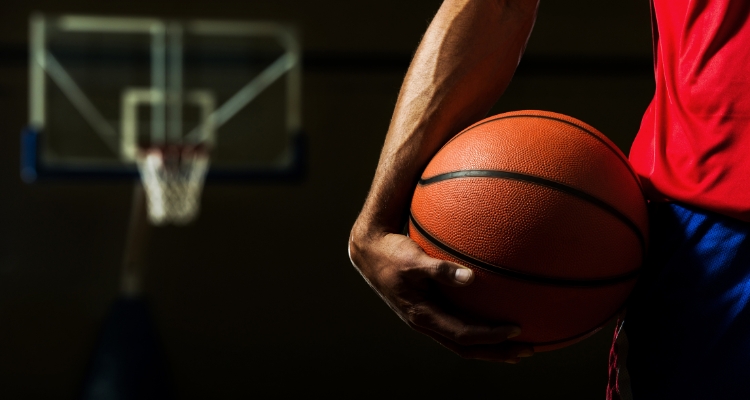 A brown-skinned arm and hand hold a basketball against a hip wearing blue athletic shorts and a red top; in the background, an out-of-focus basketball hoop can be seen.