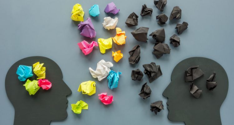 A grey background with two heads cut out of paper on top of the background. There is rainbow paper crumpled "inside" the head of the left person, with it also coming out of her mouth like positive words. The right head has black crumpled paper "inside" their head with it coming out of their mouth, like negative words.