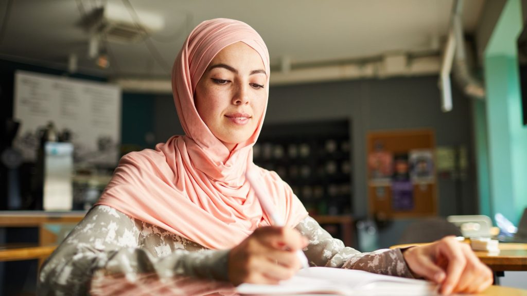 A light-skinned woman in a pink hijab writes in a notebook while sitting in a cafe.