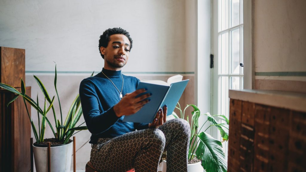 A Black person with short hair and a blue turtleneck sits in a chair reading a blue book by a window to their right. They are framed by large potted plants, and the walls around them are white.