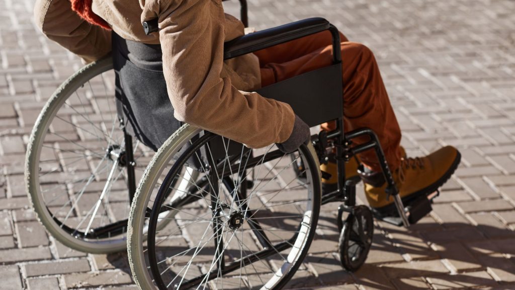 A person in a wheelchair wearing a beige jacket, orange pants, and boots is depicted at rest on a brick surface.