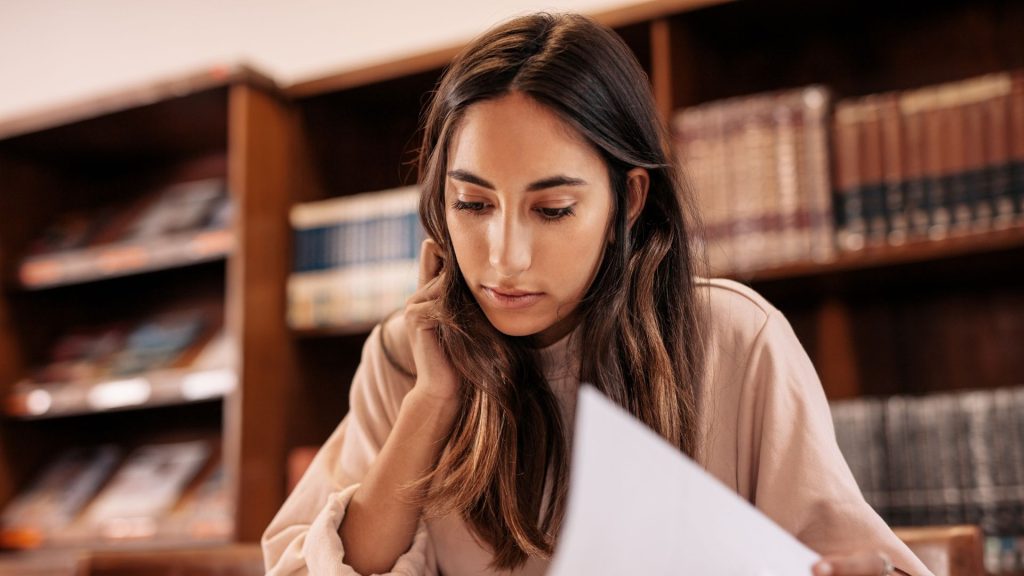 A light-skinned woman with long brown hair and a long-sleeved shirt sits at a table reading a book with library bookshelves behind her.