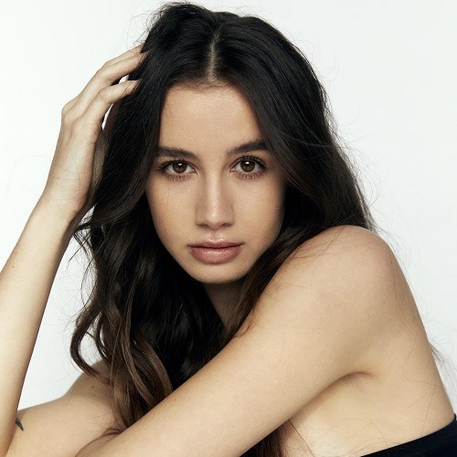 Mads Rafferty, a light-skinned young woman with long dark hair, stares at the viewer as she poses, arms leaned on a table out of frame with her right hand touching her hair. The background is white.