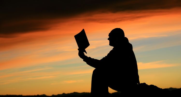 A silhouetted figure of a man sitting and reading against a sunset.