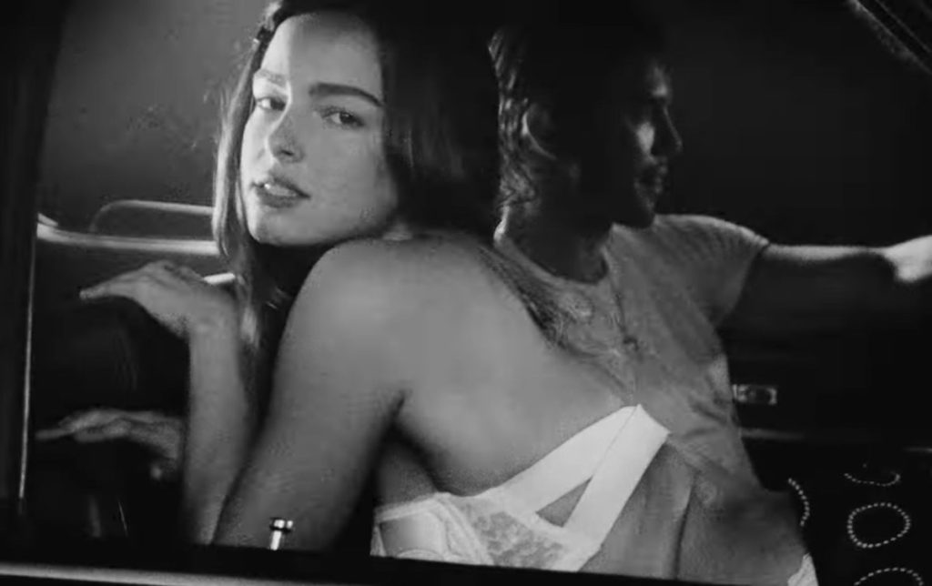 Black and white image of Addison Rae from Diet Pepsi Music Video sitting backwards on a passenger seat looking a the camera. There is a man in the driver's seat next to her.