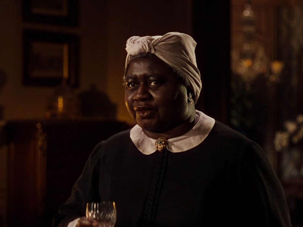 A still from the 1939 movie Gone With the Wind. A Black woman in a black and white service uniform with a white headscarf stands in a dark hall, looking concerned.