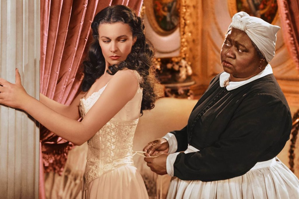 A still from the 1939 film Gone With the Wind. A young White woman braces herself against a stone column while a Black woman in a service uniform laces her corset. Behind them is an opulent Victorian bedroom with pink and gold accents.