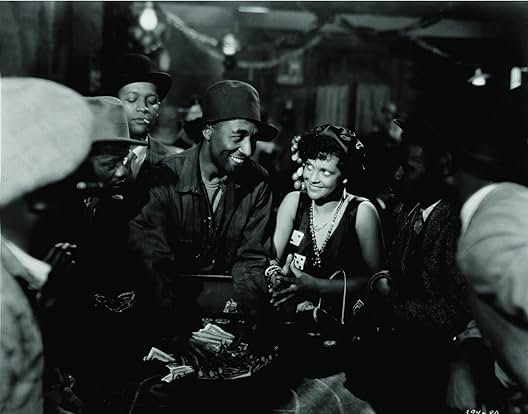 A black-and-white still from the 1929 film Hallelujah. A Black man and Black woman smile at each other in front of a casino table. Around them, other Black men watch the betting taking place.