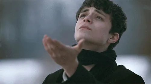 A still from Anne With an E. A young boy wearing black looks up at the sky and holds his hand out to catch fallen snow.